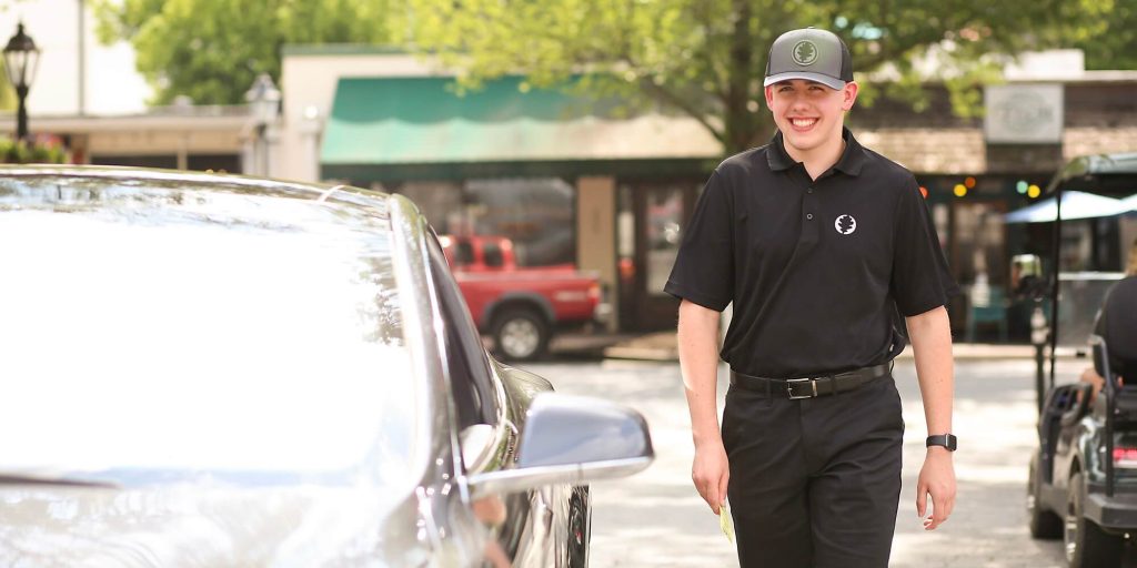Parking attendant in uniform walking toward a parked car on a tree-lined street, holding a ticket and smiling.