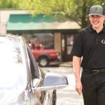 Parking attendant in uniform walking toward a parked car on a tree-lined street, holding a ticket and smiling.