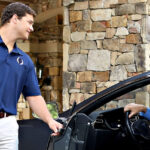 Valet attendant opening a car door and greeting a driver at a building entrance.