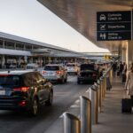 Busy airport curbside pickup and drop-off area with organized traffic flow, travelers with luggage, and multiple vehicles moving efficiently along the terminal curb.