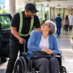 Hospital parking attendant assisting a smiling patient in a wheelchair at the medical center entrance, demonstrating compassionate, accessible patient arrival service.